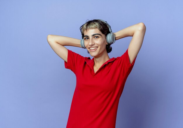 Joyful young caucasian girl with pixie haircut wearing headphones putting hands behind head isolated on purple background with copy space