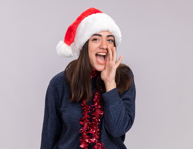 Joyful young caucasian girl with santa hat and garland around neck holds hand close to mouth calling someone 