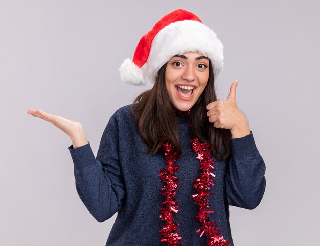 Joyful young caucasian girl with santa hat and garland around neck holds hand open and thumbs up isolated on white wall with copy space