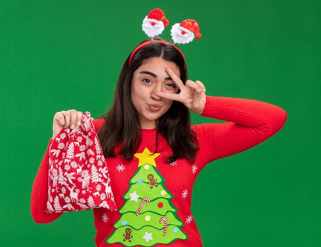 Joyful young caucasian girl with santa headband gestures victory sign and holds christmas gift bag isolated on green background with copy space