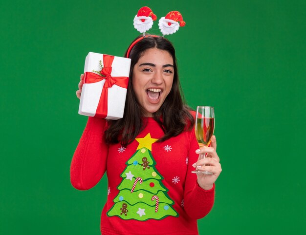 joyful young caucasian girl with santa headband holds glass of champagne and christmas gift box isolated on green background with copy space