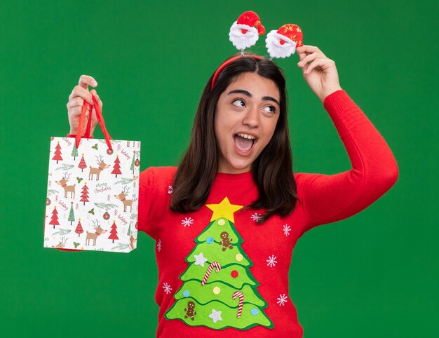 Joyful young caucasian girl with santa headband holds paper gift bag looking at side isolated on green background with copy space