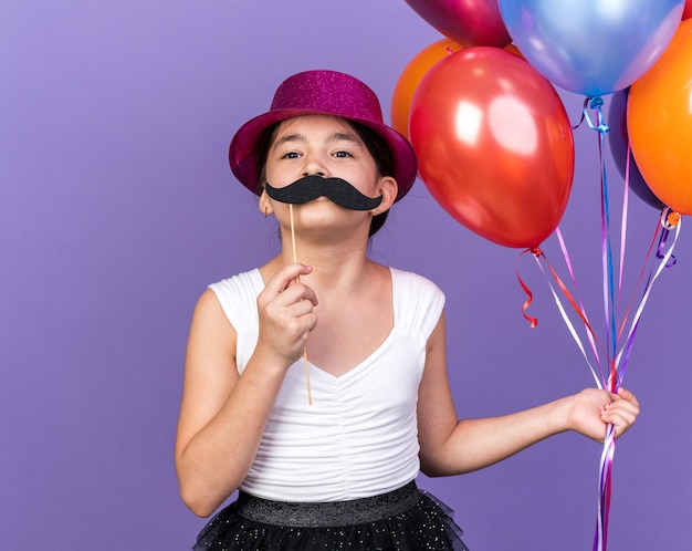 joyful young caucasian girl with violet party hat holding helium balloons and fake mustache on stick isolated on purple wall with copy space