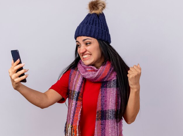 Joyful young caucasian ill girl wearing winter hat and scarf holding and looking at mobile phone doing yes gesture isolated on white wall with copy space