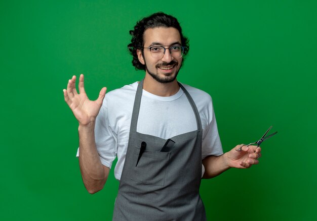 Joyful young caucasian male barber wearing glasses and wavy hair band in uniform holding scissors and keeping hand in air isolated on green background