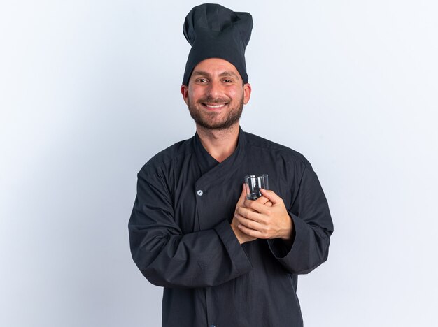 Joyful young caucasian male cook in chef uniform and cap holding glass of water in front of heart 