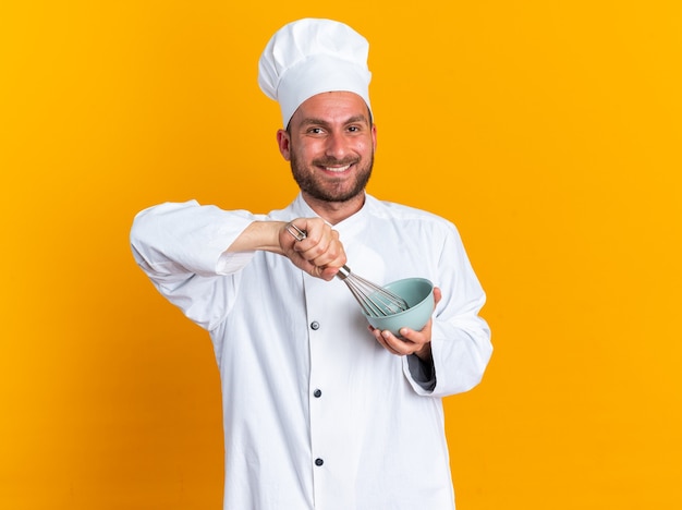 Joyful young caucasian male cook in chef uniform and cap whisking eggs in bowl looking at camera isolated on orange wall with copy space