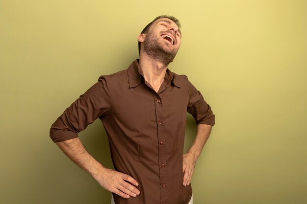 Joyful young caucasian man keeping hands on waist laughing with closed eyes isolated on olive green background with copy space