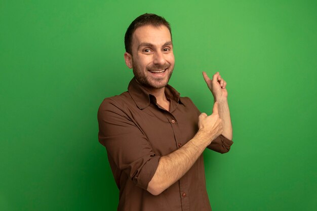 Joyful young caucasian man looking at camera pointing behind isolated on green background with copy space