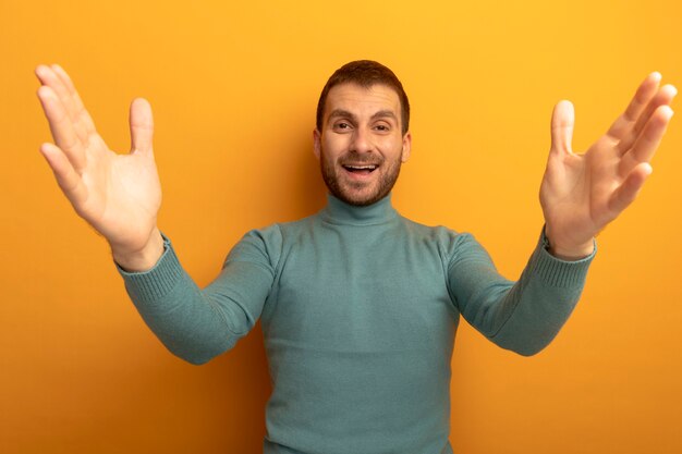 Joyful young caucasian man looking at camera spreading hands isolated on orange background