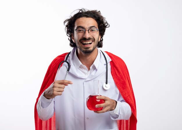 Joyful young caucasian man in optical glasses wearing doctor uniform with red cloak and with stethoscope around neck holds and points at red chemical liquid in glass flask 