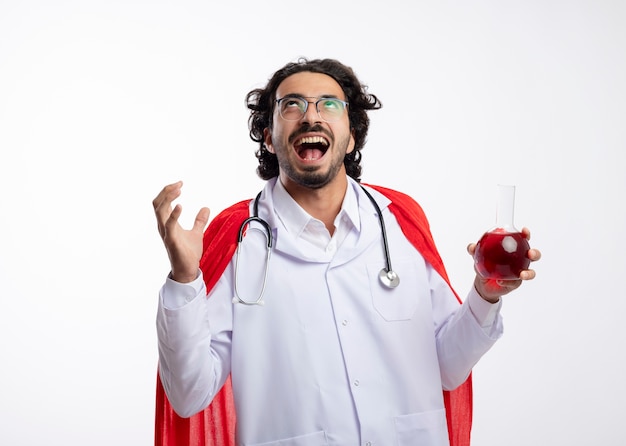 Foto gratuita gioioso giovane uomo caucasico in vetri ottici che indossa l'uniforme del medico con mantello rosso e con lo stetoscopio intorno al collo sta con la mano alzata e tiene il liquido chimico rosso nel pallone di vetro