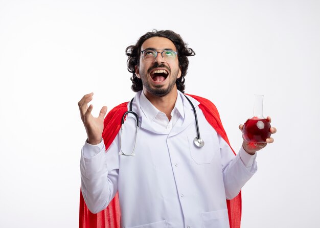 Joyful young caucasian man in optical glasses wearing doctor uniform with red cloak and with stethoscope around neck stands with raised hand and holds red chemical liquid in glass flask