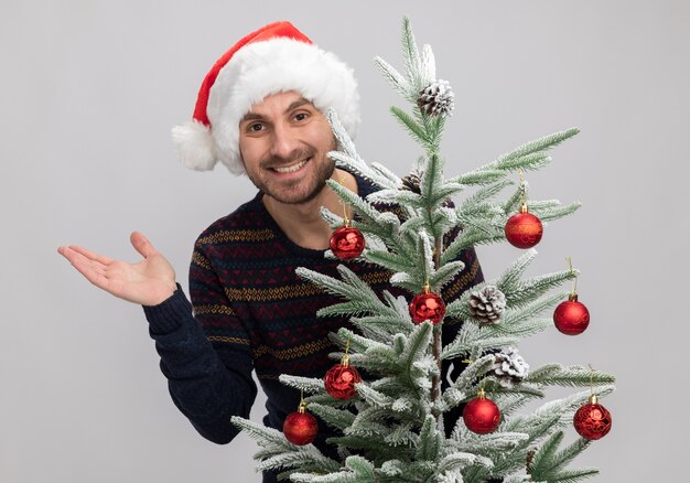 Joyful young caucasian man wearing christmas hat standing behind christmas tree looking at camera showing empty hand isolated on white background