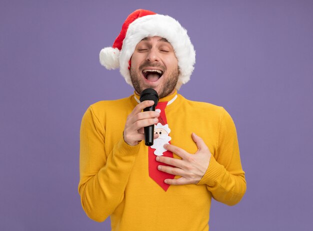크리스마스 모자를 입고 즐거운 젊은 백인 남자와 보라색 배경에 고립 된 가슴에 손을 유지 닫힌 눈으로 마이크 노래를 들고 넥타이