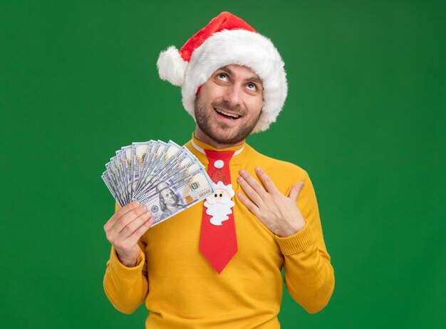 Joyful young caucasian man wearing christmas hat and tie holding money putting hand on chest looking up isolated on green wall with copy space