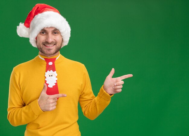 Joyful young caucasian man wearing christmas hat and tie looking at camera pointing at side isolated on green background