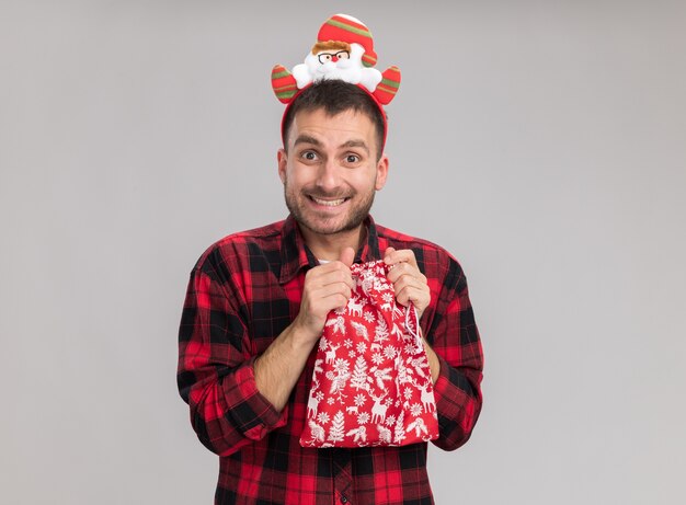 Joyful young caucasian man wearing christmas headband holding christmas sack looking at camera isolated on white background