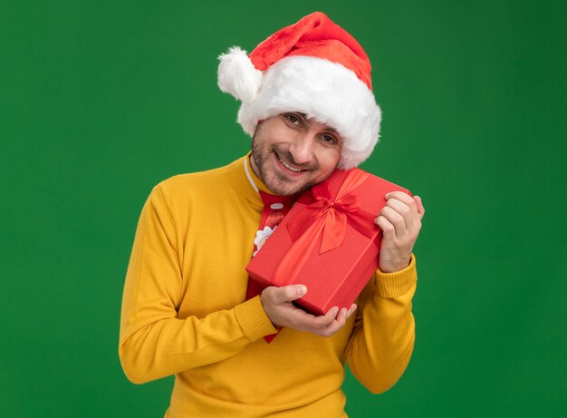 Joyful young caucasian man wearing christmas tie and hat holding gift package looking at camera isolated on green background