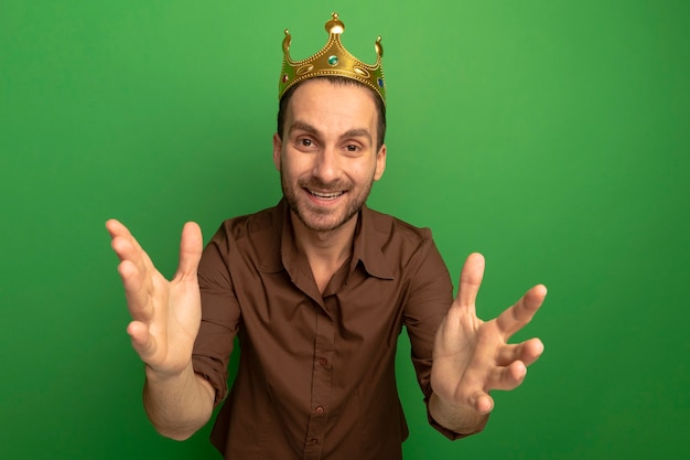 Free photo joyful young caucasian man wearing crown looking at camera stretching out hands towards camera isolated on green background