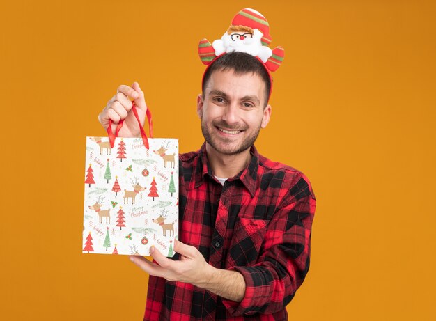 Joyful young caucasian man wearing santa claus headband holding christmas gift bag looking at camera isolated on orange background