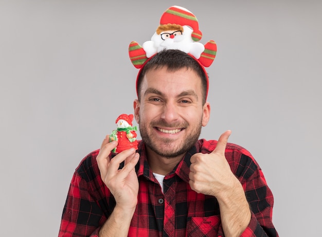 雪だるまのクリスマス飾りを保持しているサンタクロースのヘッドバンドを身に着けているうれしそうな若い白人男性が白い背景で隔離の親指を上に表示しているカメラを見て