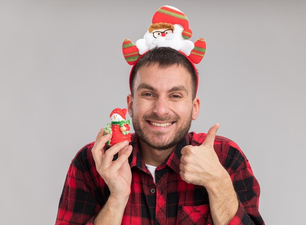 雪だるまのクリスマス飾りを保持しているサンタクロースのヘッドバンドを身に着けているうれしそうな若い白人男性は、白い壁に分離された親指を示しています