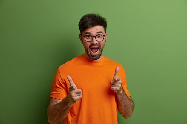 Joyful young Caucasian man with beard makes finger gun gesture, points at you, selects someone, wears optical glasses and orange t shirt, makes choice, isolated on green  wall. You my bro