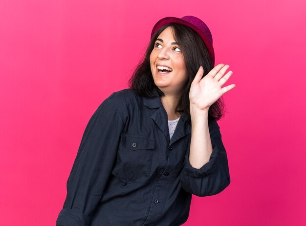 Joyful young caucasian party woman wearing party hat looking up doing hi gesture isolated on pink wall