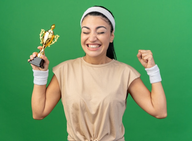 Радостная молодая кавказская спортивная девушка с повязкой на голову и браслетами, держащая кубок победителя, делает жест `` да '' с закрытыми глазами, изолированными на зеленой стене