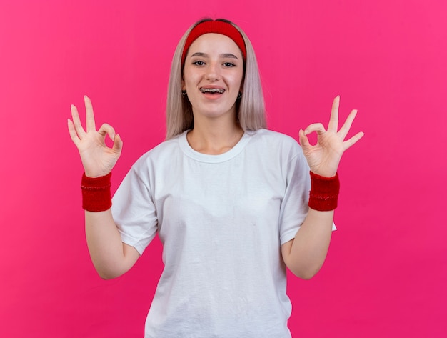 Joyful young caucasian sporty girl with braces wearing headband and wristbands gestures ok hand sign with two hands 