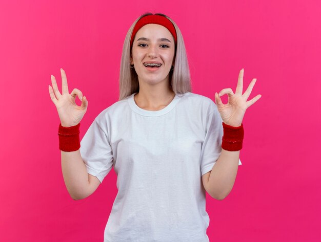Радостная молодая кавказская спортивная девушка с подтяжками, носящая повязку на голову и браслеты, жестами показывает знак рукой двумя руками