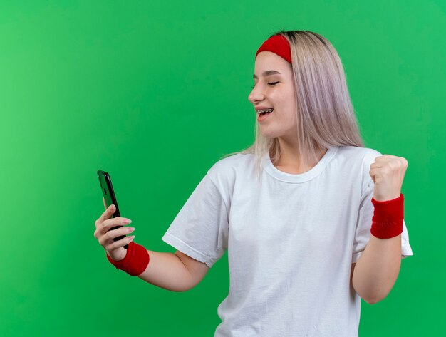 Радостная молодая кавказская спортивная девушка с подтяжками, носящая повязку на голову и браслеты держит кулак и смотрит на телефон