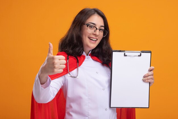 Joyful young caucasian superhero girl in red cape wearing doctor uniform and stethoscope with glasses showing clipboard to camera showing thumb up 