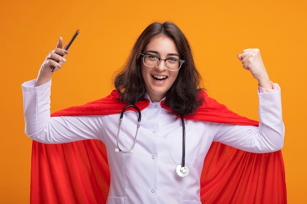 Joyful young caucasian superhero girl wearing doctor uniform and stethoscope with glasses holding mobile phone  doing yes gesture isolated on wall