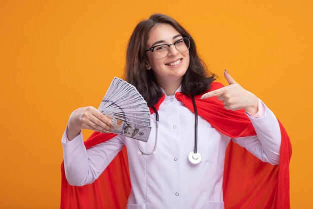 Joyful young caucasian superhero girl wearing doctor uniform and stethoscope with glasses holding and pointing at money  isolated on wall