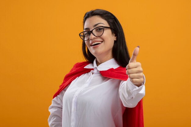 Joyful young caucasian superhero girl wearing glasses standing in profile view looking at camera showing thumb up isolated on orange background with copy space