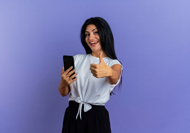 Joyful young caucasian woman holds phone and thumbs up looking at camera