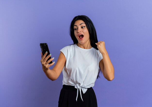 Joyful young caucasian woman keeps fist holding and looking at phone