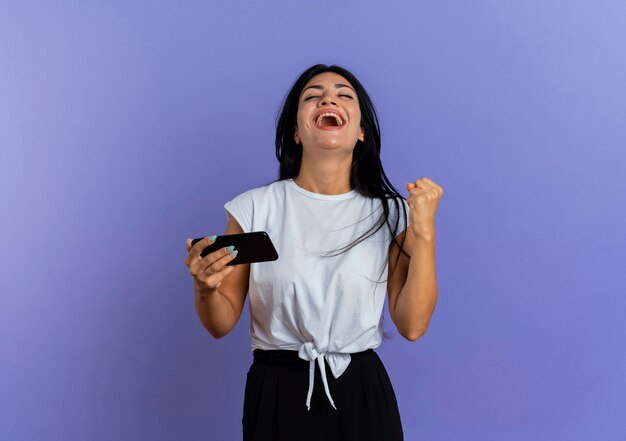 Joyful young caucasian woman keeps fist and holds phone looking up