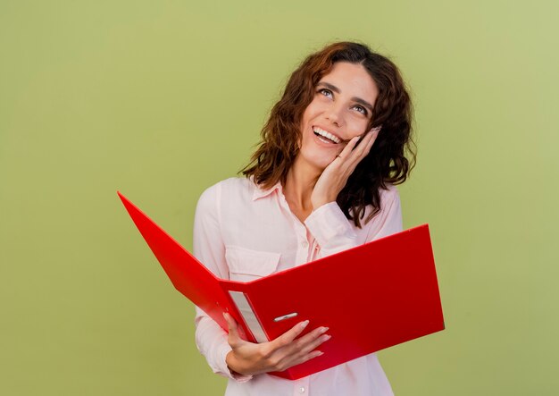 Joyful young caucasian woman puts hand on face looking up and holding file folder isolated on green background with copy space