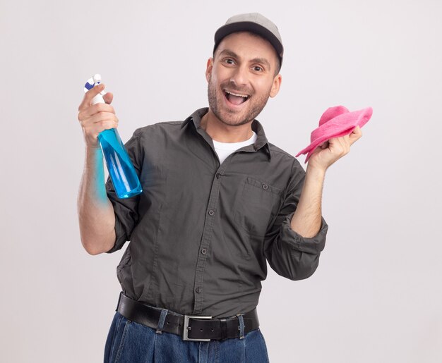 Joyful young cleaning man wearing casual clothes and cap holding cleaning spray and rag  happy and excited standing over white wall