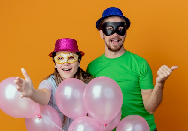 Gioiosa giovane coppia in piedi con palloncini ad elio ragazza con cappello rosa che indossa una maschera per gli occhi in maschera punta in avanti con la mano bell'uomo con cappello blu che indossa una maschera per gli occhi in maschera punta a lato