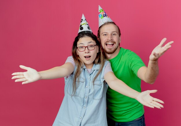 파티 모자를 쓰고 즐거운 젊은 부부는 분홍색 벽에 고립 된 손을 올리는 모습