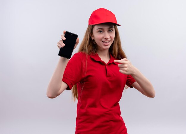 Joyful young delivery girl in red uniform holding and pointing at mobile phone on isolated white space