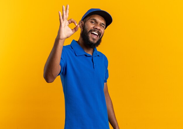 joyful young delivery man blinks his eye gesturing ok sign isolated on orange wall with copy space