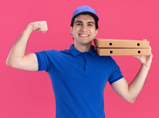 Joyful young delivery man in blue uniform and cap holding pizza packages on shoulder looking at front doing strong gesture isolated on pink wall