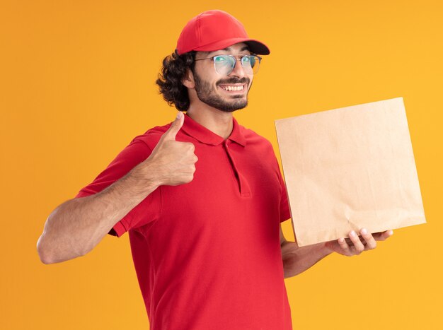 赤い制服を着たうれしそうな若い配達人とオレンジ色の壁に分離された親指を上に表示して正面を見て紙のパッケージを保持している眼鏡をかけている