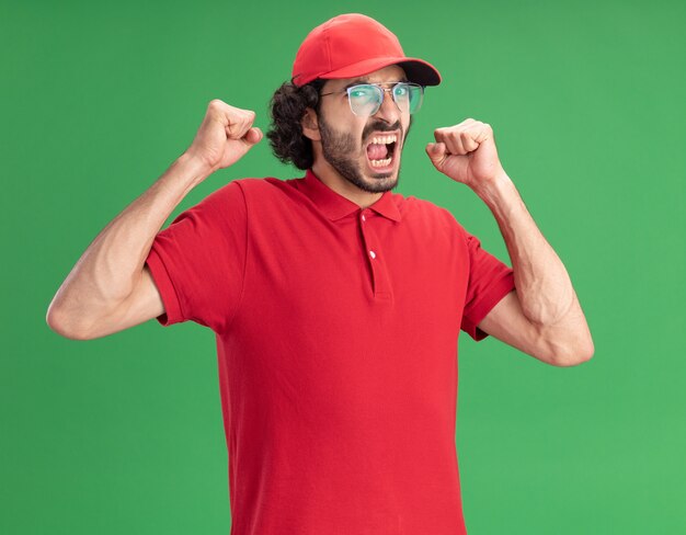 Joyful young delivery man in red uniform and cap wearing glasses looking at front doing yes gesture isolated on green wall