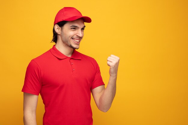 joyful young delivery man wearing uniform and cap looking at side showing yes gesture isolated on yellow background with copy space